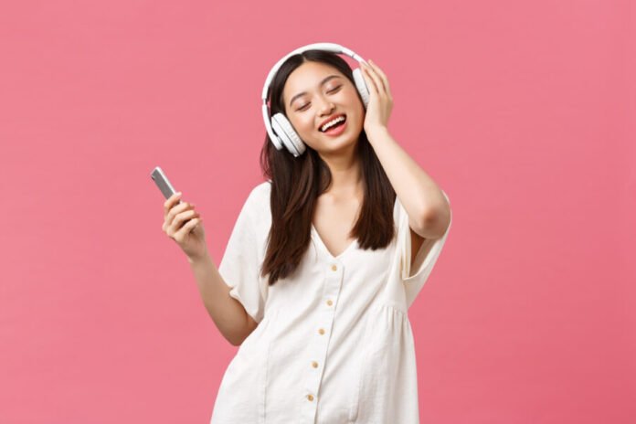Beauty, people emotions and technology concept. Carefree and pretty asian girl enjoying music in wireless headphones, close eyes and smiling listening favorite song, holding smartphone and dancing
